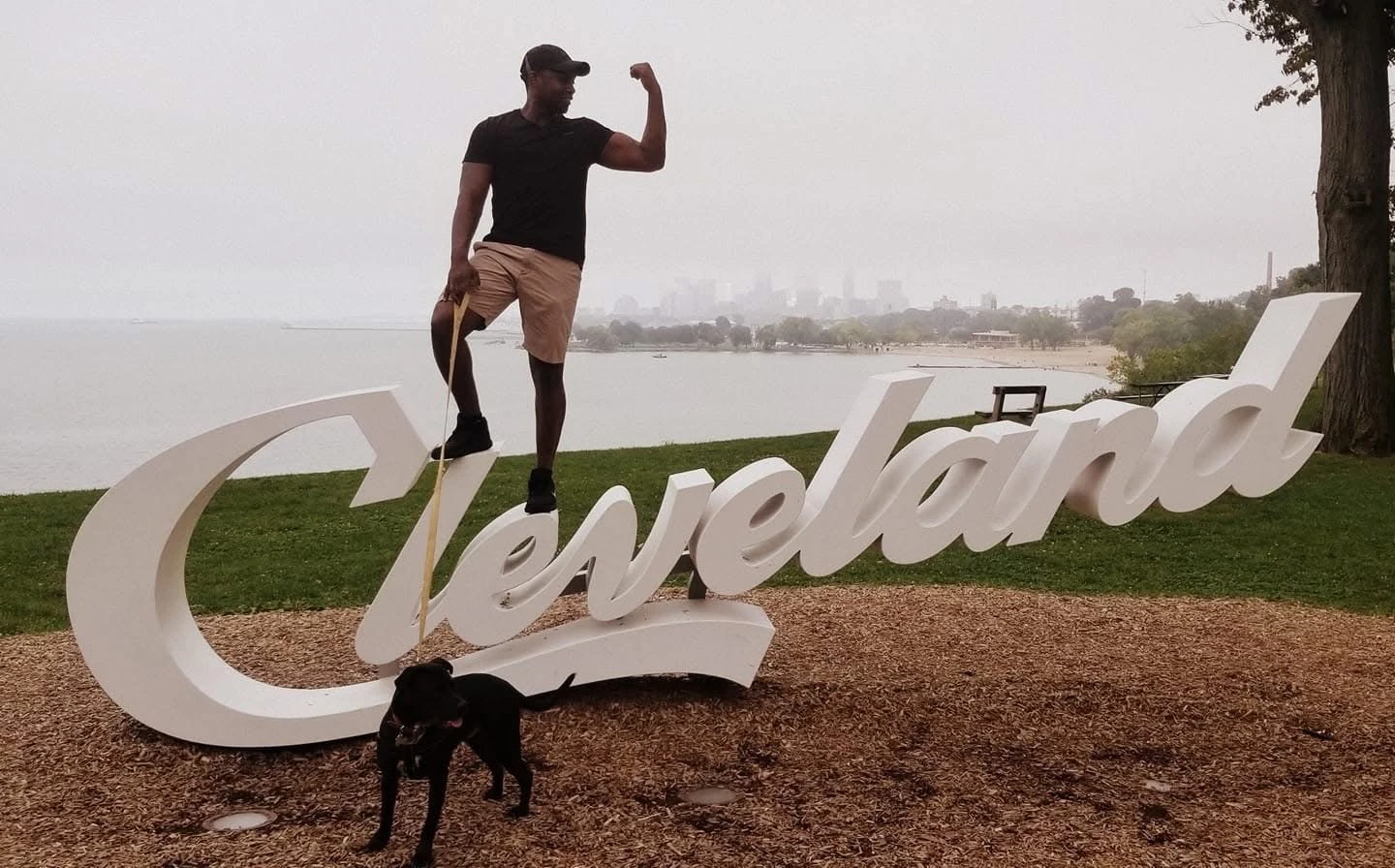 Matthew flexing beside the Cleveland sign by the lake, with a dog in the foreground
